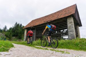 Two mountain bikers riding past a traditional wooden barn on the Idrija MTB route in Slovenia